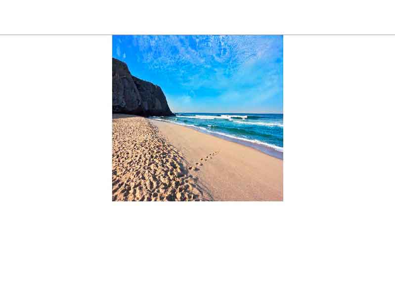Ein sonniger Sandstrand mit Fußspuren im Sand, einer felsigen Klippe und blauem Himmel am Meer
