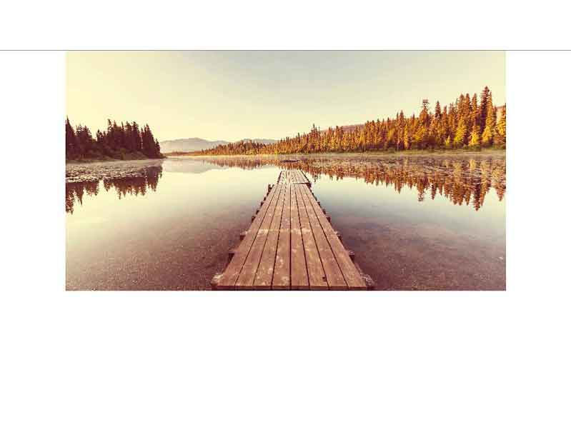 Holzsteg über einem ruhigen See bei Sonnenuntergang Ein langer Holzsteg führt über einen ruhigen See, umgeben von einem Wald und Bergen im Hintergrund, bei Sonnenuntergang.